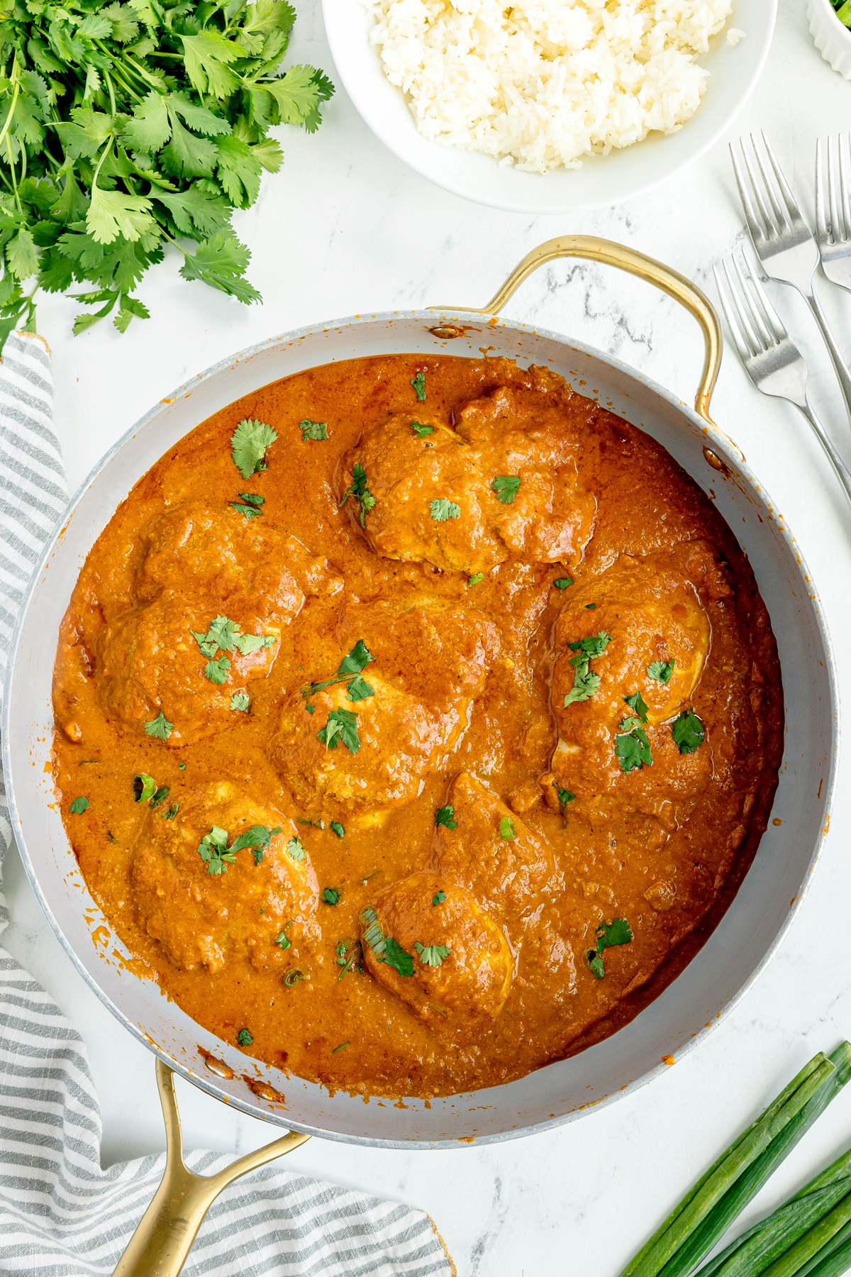A pan filled with Butter Chicken in a thick orange curry sauce, garnished with cilantro, sits on a white surface next to rice, fresh herbs, and forks.