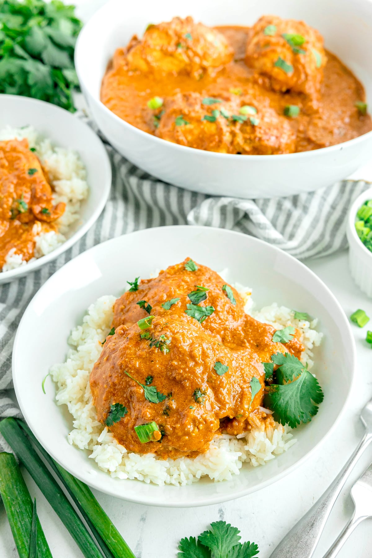 A bowl of white rice topped with saucy Butter Chicken curry, garnished with chopped cilantro, sits in the foreground with a larger serving dish and extra garnishes in the background.