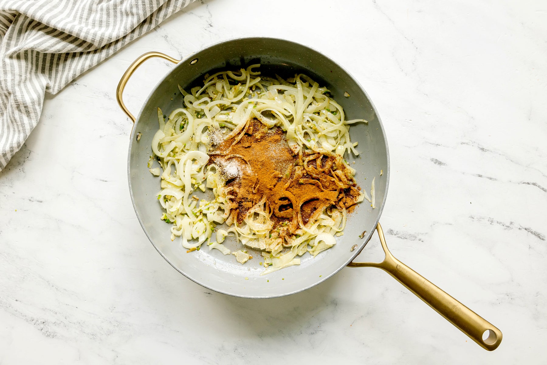 Sliced onions cooking in a gray pan with garam masala, cinnamon, salt, and pepper sprinkled on top, placed on a white marble countertop beside a striped cloth for Butter Chicken Recipe