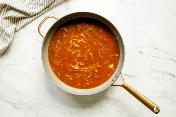 Next step in preparing Butter Chicken is to add tomato puree and chicken stock in a pot. Visible onion slices sits on a white marble surface next to a striped cloth.