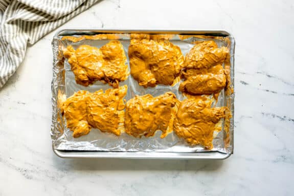 Next step in preparing Butter Chicken is to put six pieces of raw chicken coated in a yellow marinade on a foil-lined baking sheet, ready for cooking. A striped towel is nearby on a marble countertop.