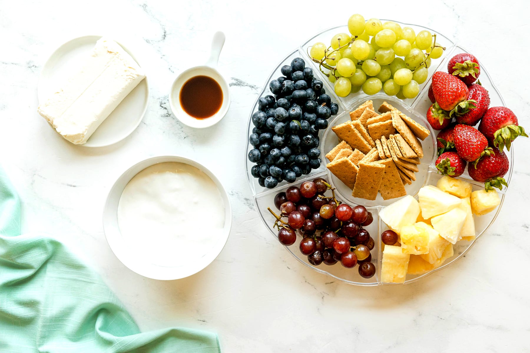 A platter with ingredients for Easy Fruit Dip are the following: cream cheese, marshmallow creme and vanilla extract, with fresh fruits such as strawberries, apple slices, grapes, pineapple, blueberries, kiwi and melon.