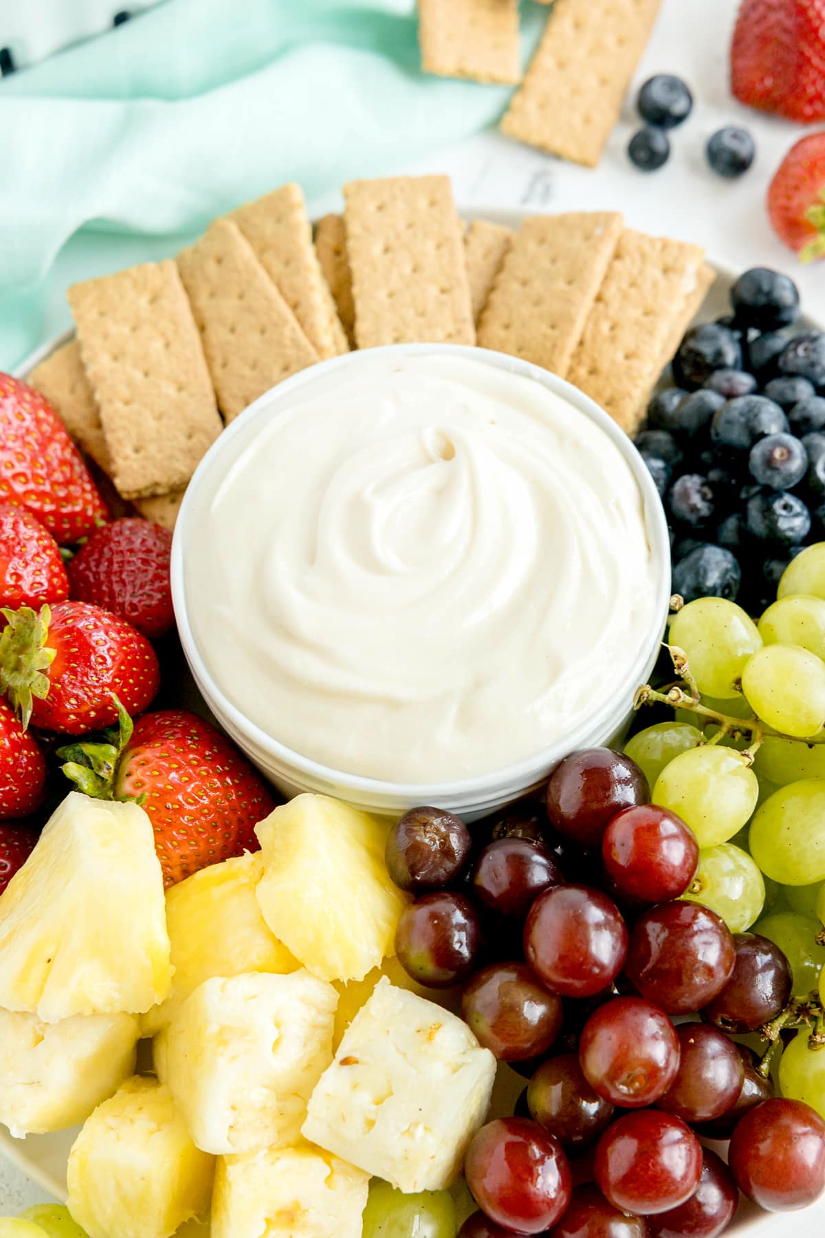 A platter with strawberries, pineapple, red and green grapes, blueberries, graham crackers, and a bowl of creamy dip in the center.