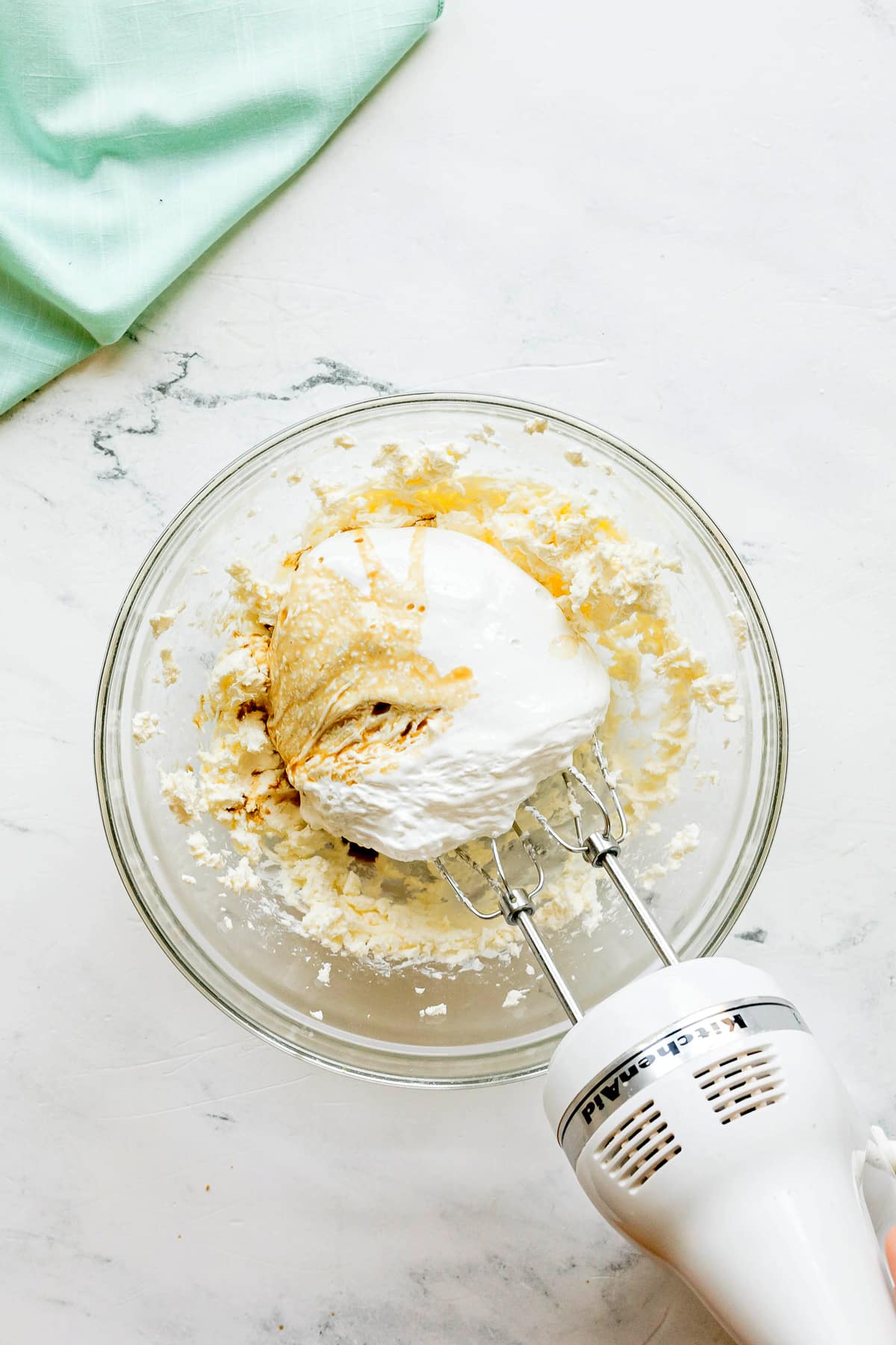 A hand mixer blending whipped ingredients for Easy Fruit Dip includes marshmallow creme, vanilla extract and orange juice, in a clear glass bowl on a white countertop.