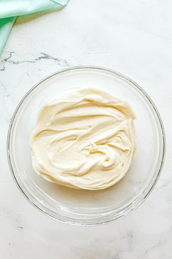 A glass bowl filled with smooth, creamy white frosting for Easy Fruit Dip on a light marble surface.