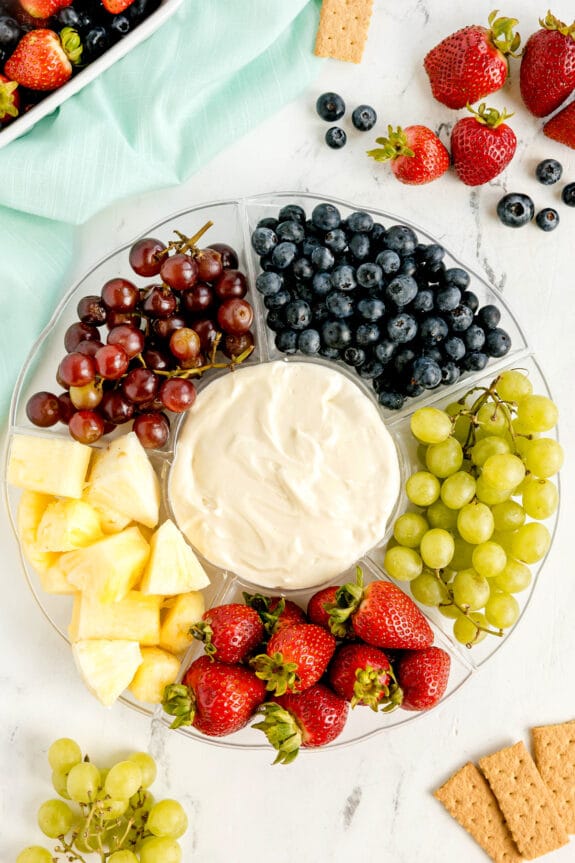A round platter with sections of grapes, blueberries, pineapple, and strawberries arranged around a central bowl of Fruit Dip, placed on a white surface with extra fruit and crackers nearby.