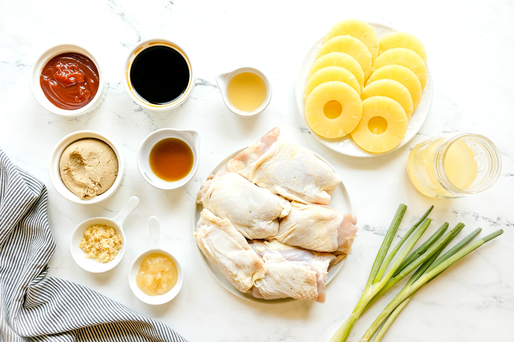Huli Huli Chicken ingredients on a white counter, are the following: canned pineapple rings, ketchup, soy sauce, brown sugar, cider vinegar, minced ginger, minced garlic, sesame oil, boin-in, skin-on chicken thighs and scallions.