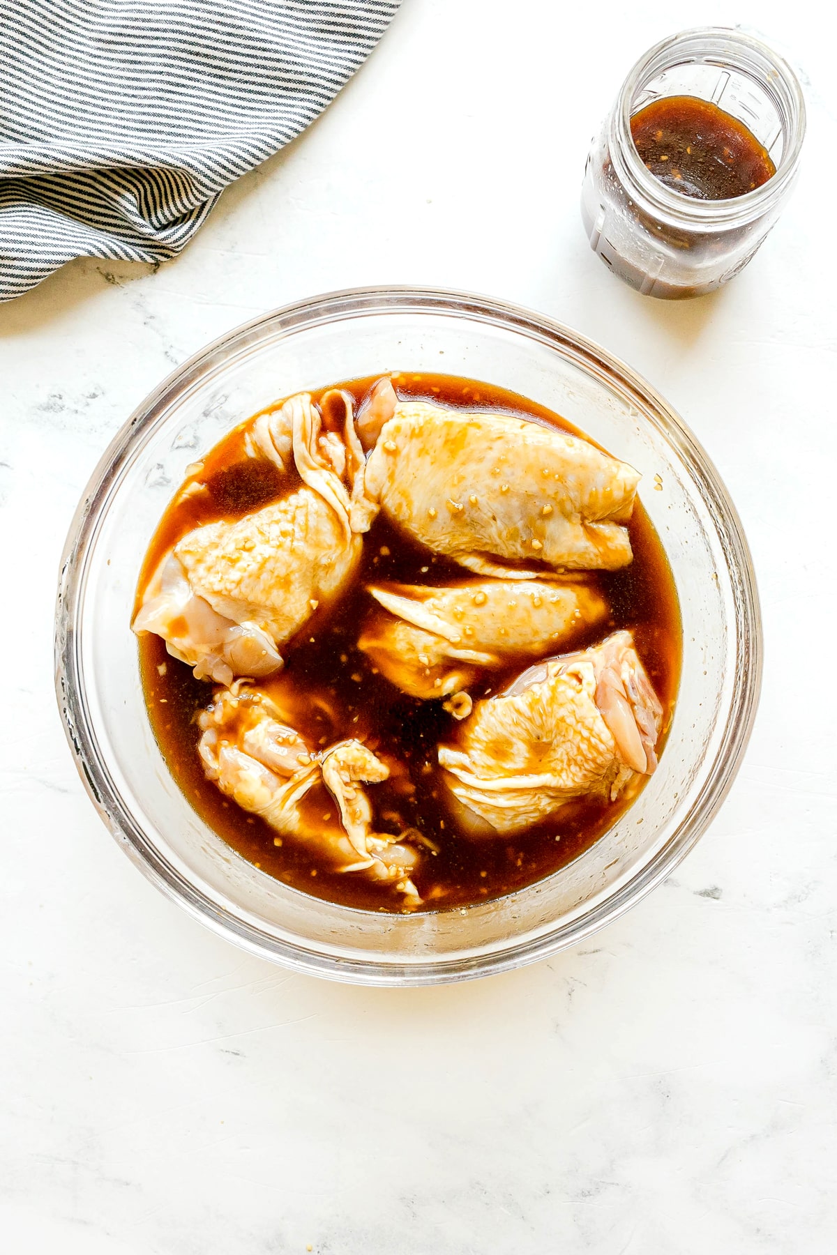 The next step in preparing Huli Huli Chicken is to add raw chicken pieces in a bowl of dark marinate sauce, with a jar of extra marinade nearby on a white surface.