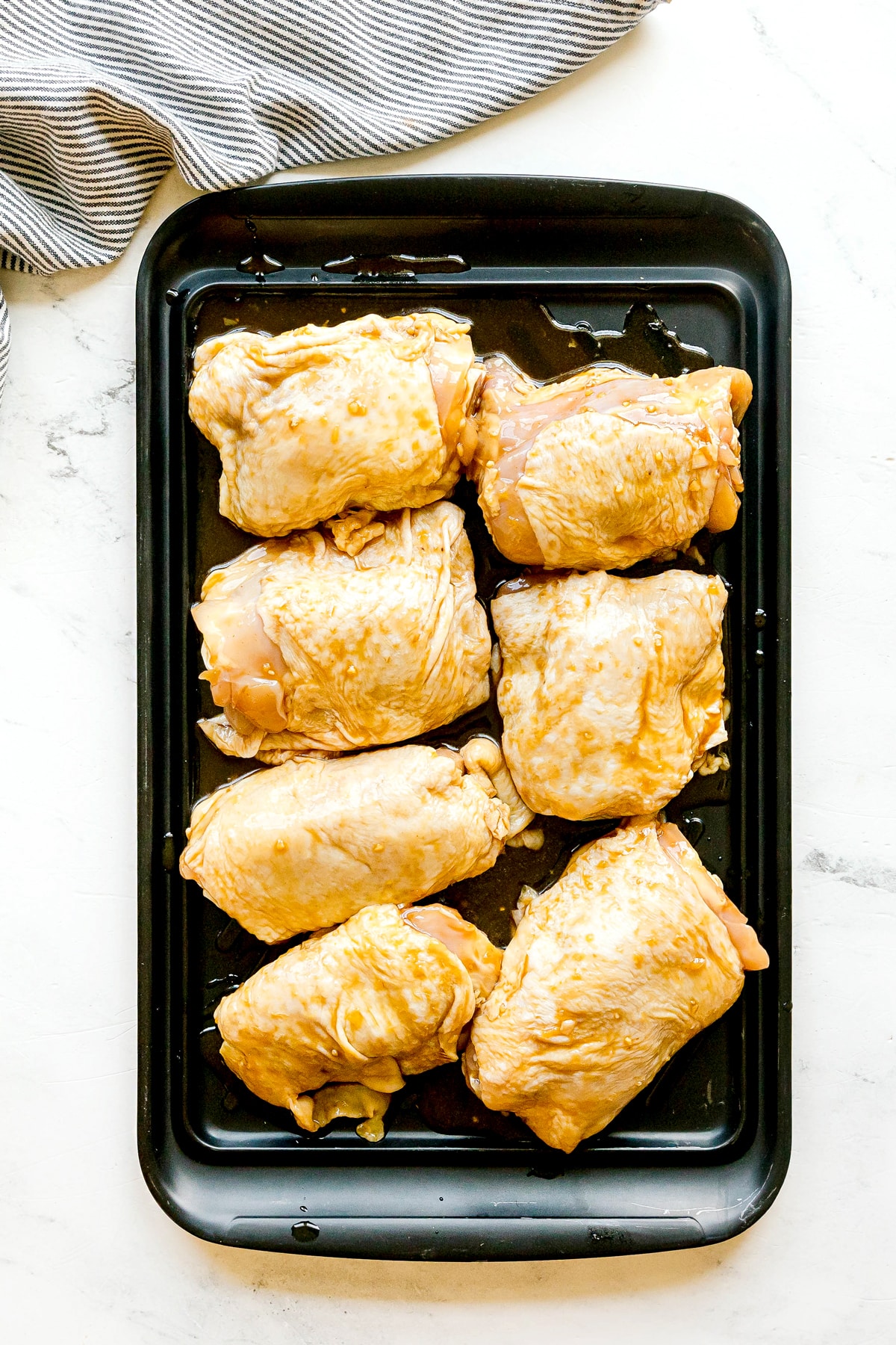 A baking tray with eight marinated raw chicken thighs for Huli Huli Chicken recipe, arranged on it, set on a white surface next to a striped cloth.