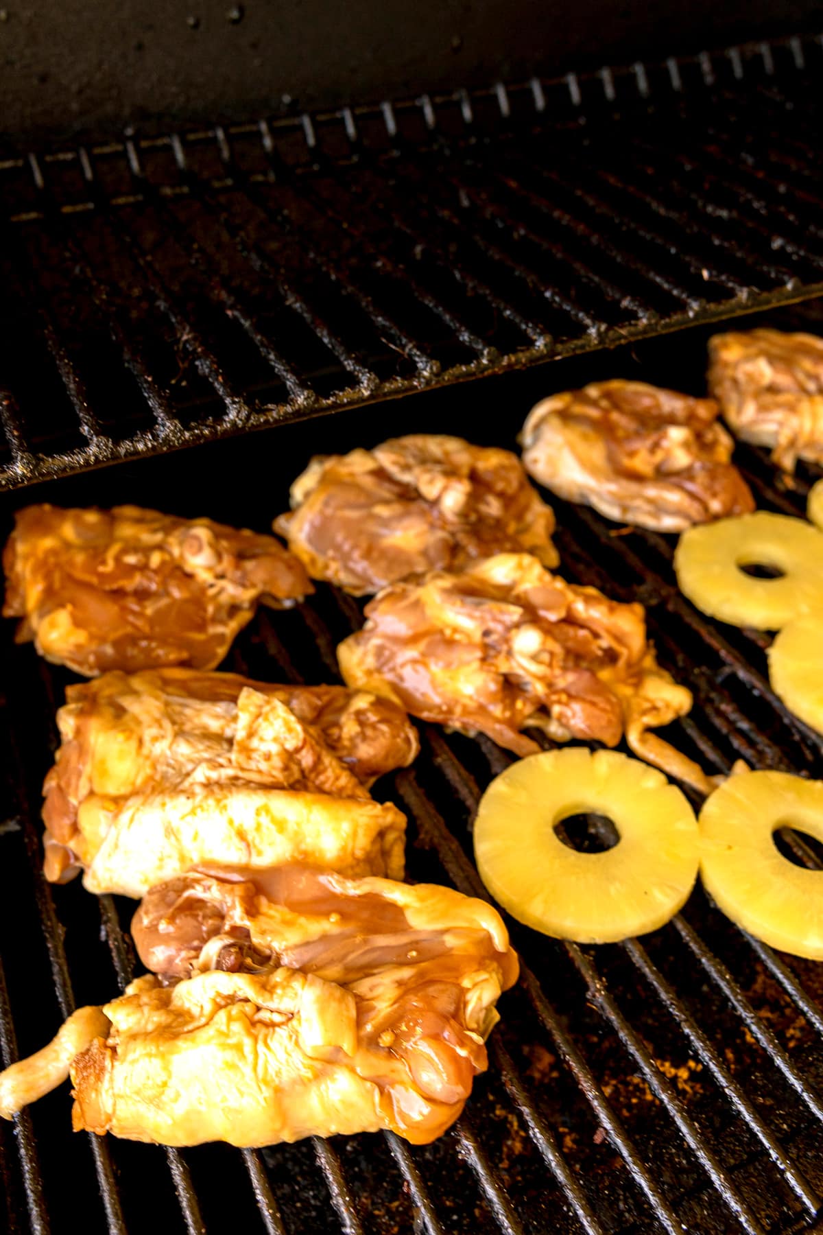 Chicken pieces and pineapple rings cooking on a grill with dark grates. This is one of the steps in preparing Huli Huli Chicken
