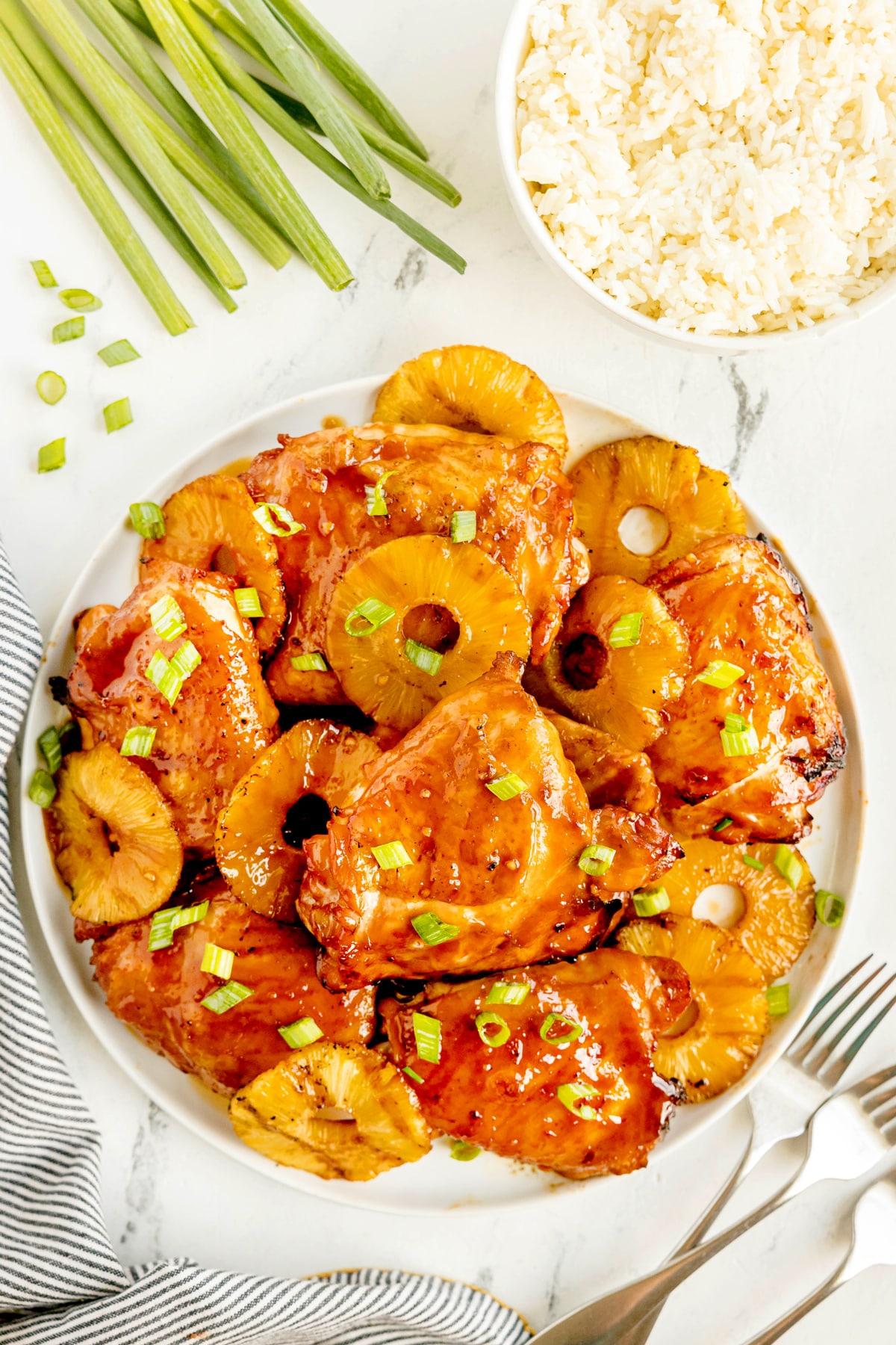 Plate of glazed Huli Huli Chicken with chopped green onions, served next to a bowl of white rice and green onions on a marble surface.