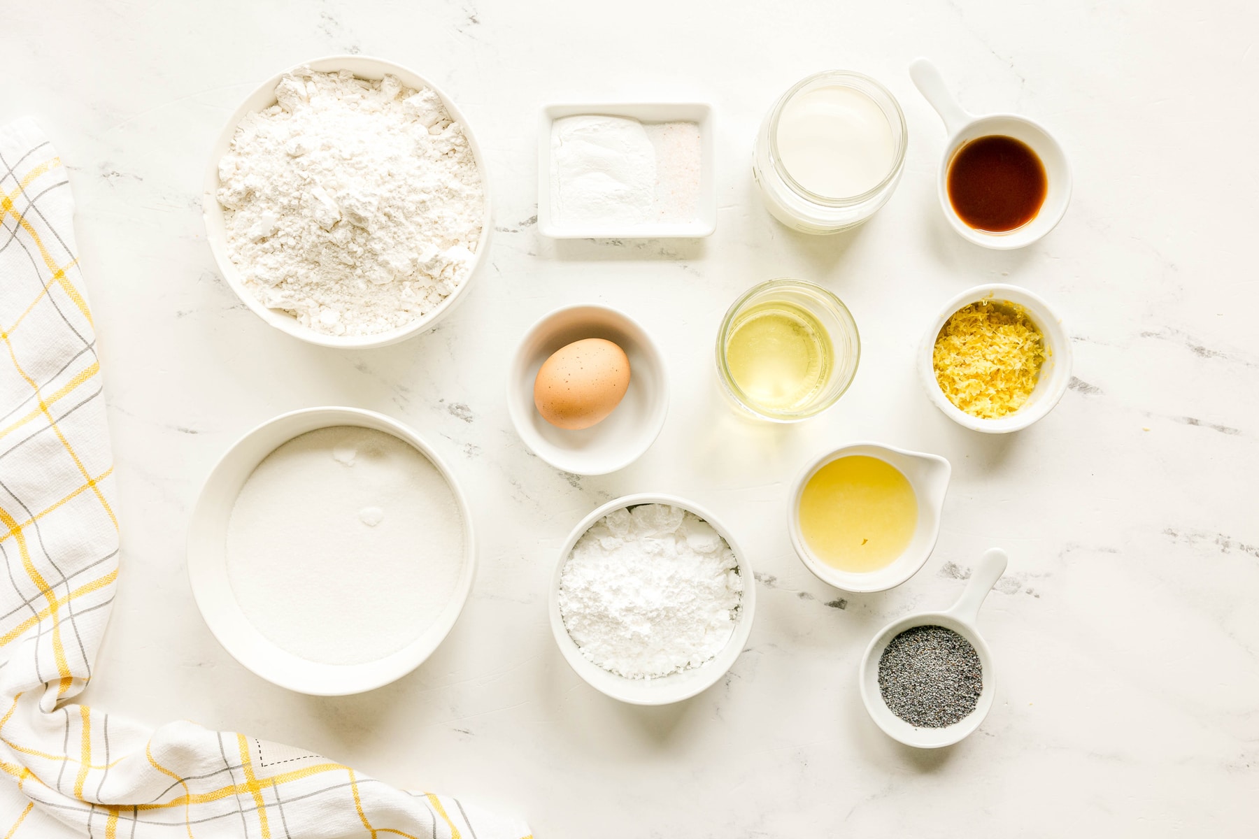 Assorted baking ingredients for Lemon Poppy Seed Bread include all-purpose flour, granulated sugar, baking powder, salt, egg, milk, vegetable oil, vanilla extract, lemon zest, lemon juice, poppy seeds, powdered sugar, lemon juice are arranged on a white surface.