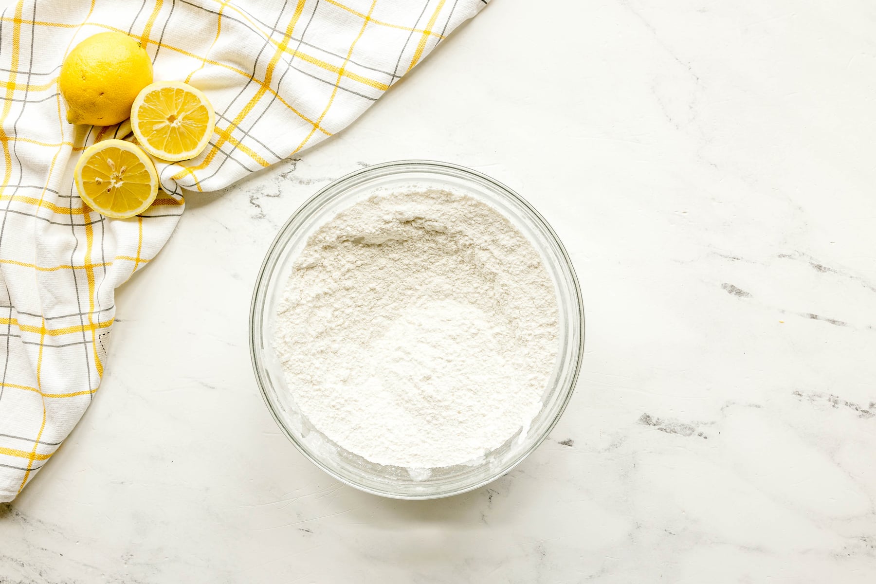 A glass bowl filled with flour, sugar, baking powder, and salt sits on a white marble surface next to a cut lemon and a yellow checkered cloth for Lemon Poppy Seed Bread.