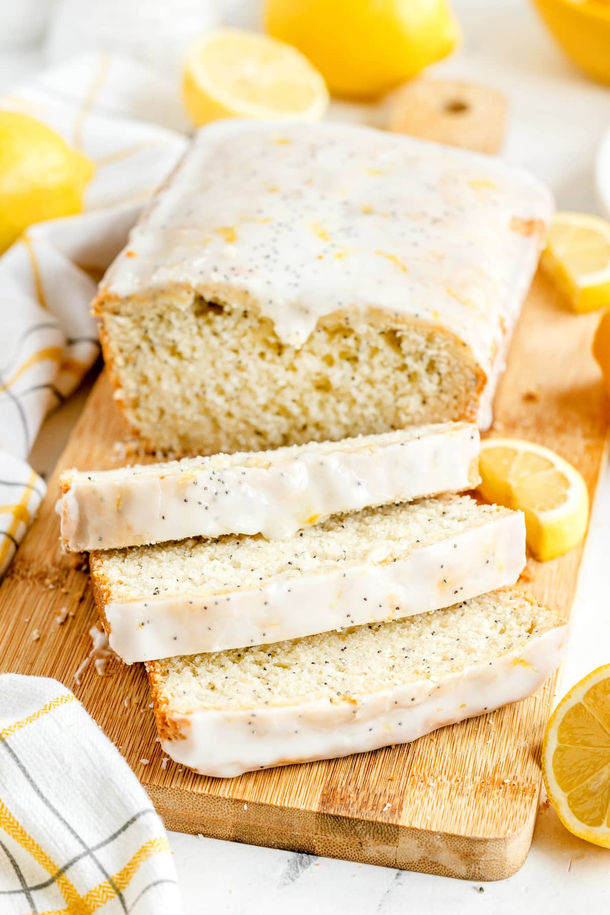 A loaf of Lemon Poppy Seed Bread with icing, partially sliced, sits on a wooden cutting board with fresh lemon halves nearby.