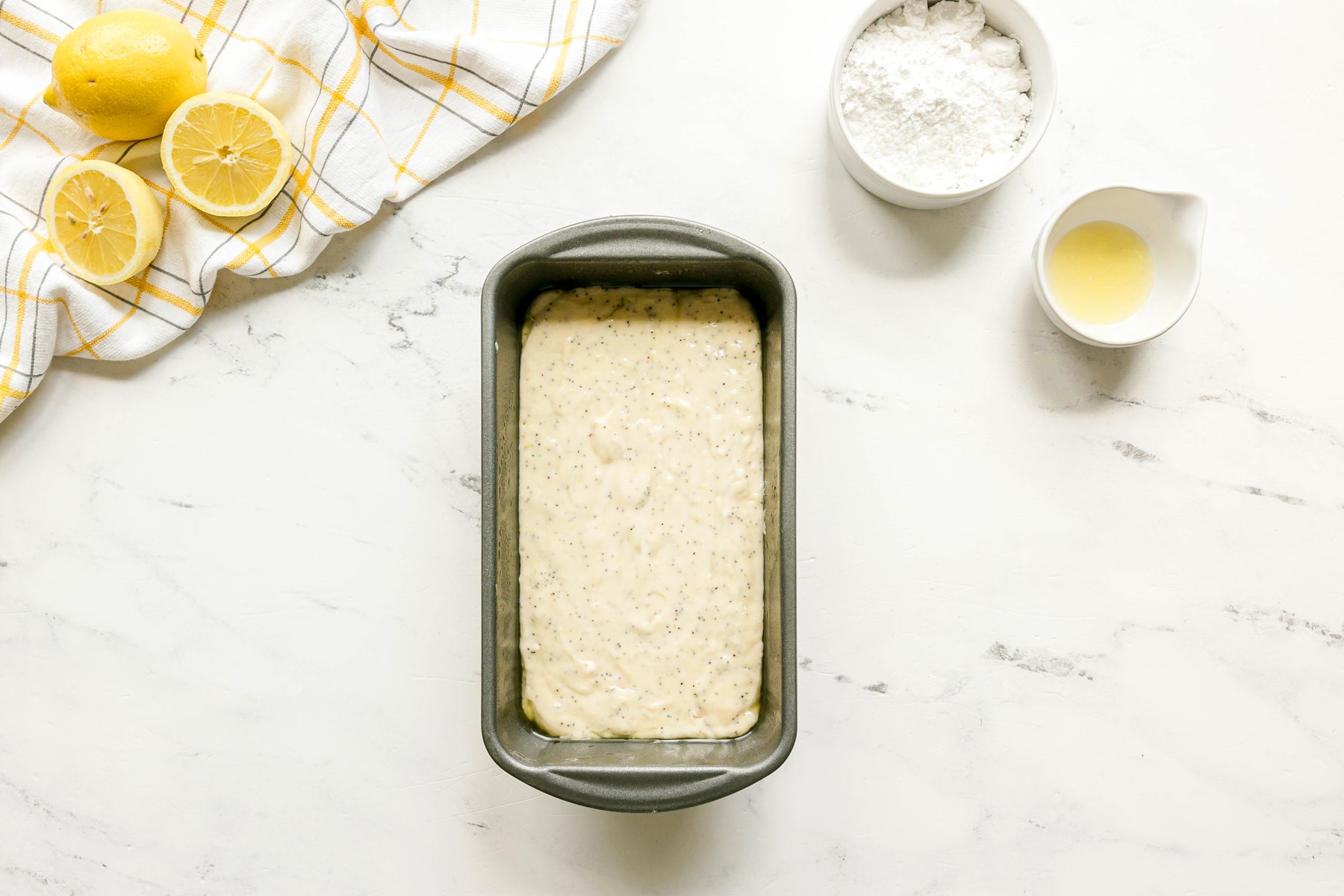 A loaf pan filled with unbaked lemon poppy seed batter sits on a white countertop next to sliced lemons, a bowl of powdered sugar, a small cup of lemon juice, and a checkered towel. This is the next step in preparing Lemon Poppy Seed Bread