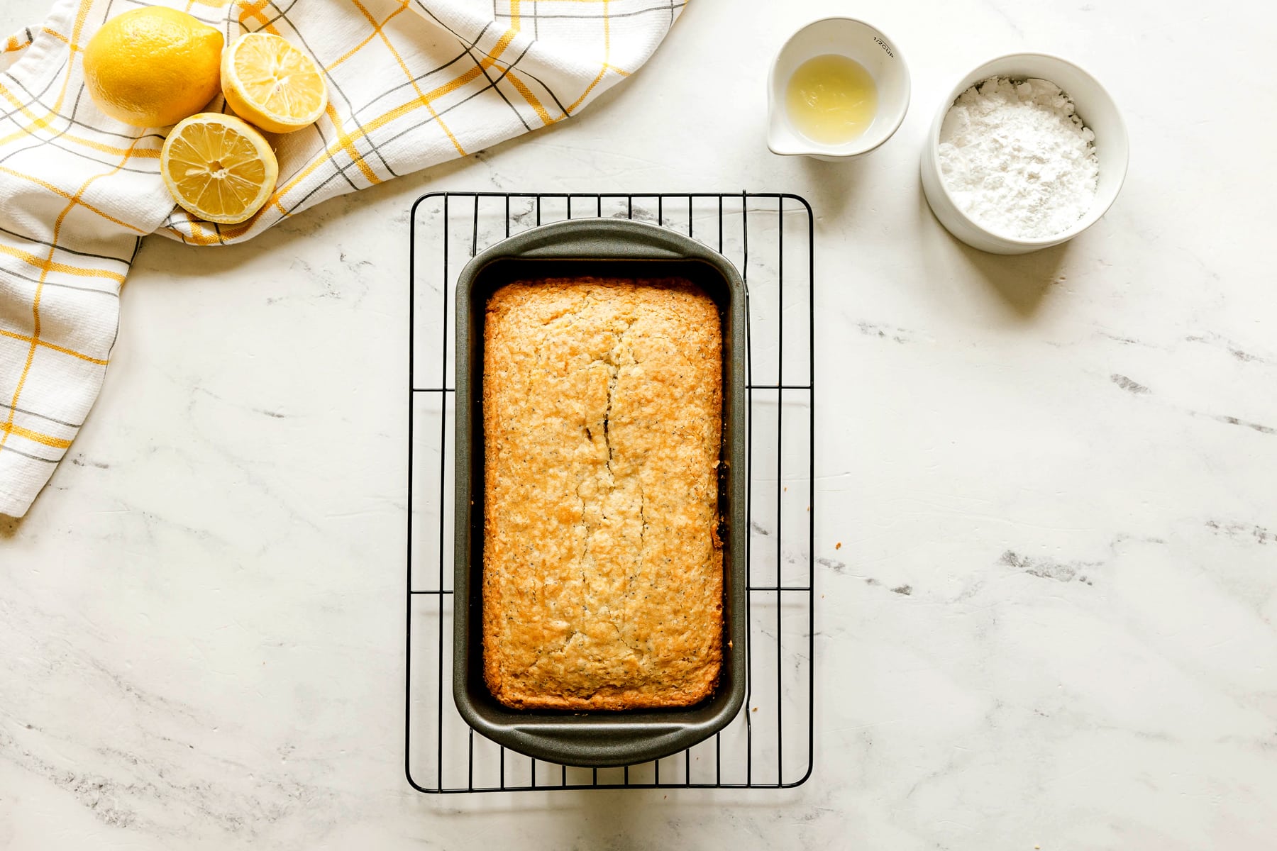 A loaf of Lemon Poppy Seed Bread on a cooling rack, with lemons, a bowl of powdered sugar, and a small dish of lemon juice on a white countertop.