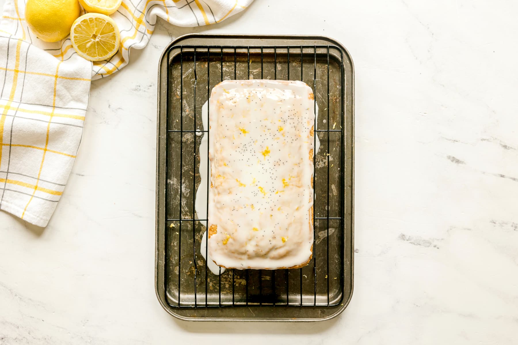 A glazed Lemon Poppy Seed Bread with lemon zest sits on a wire rack over a baking tray. A kitchen towel and sliced lemons are placed in the top left corner.