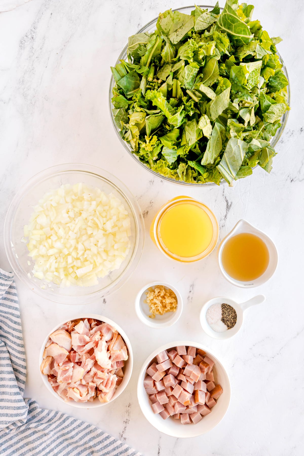 Ingredients for Southern Collard Greens are seen on bowls of hickory-smoked bacon, sweet onions, smoked ham, garlic cloves, chicken broth, fresh collard greens, apple cider vinegar, sugar, salt, and black pepper. They are arranged on a white marble surface.