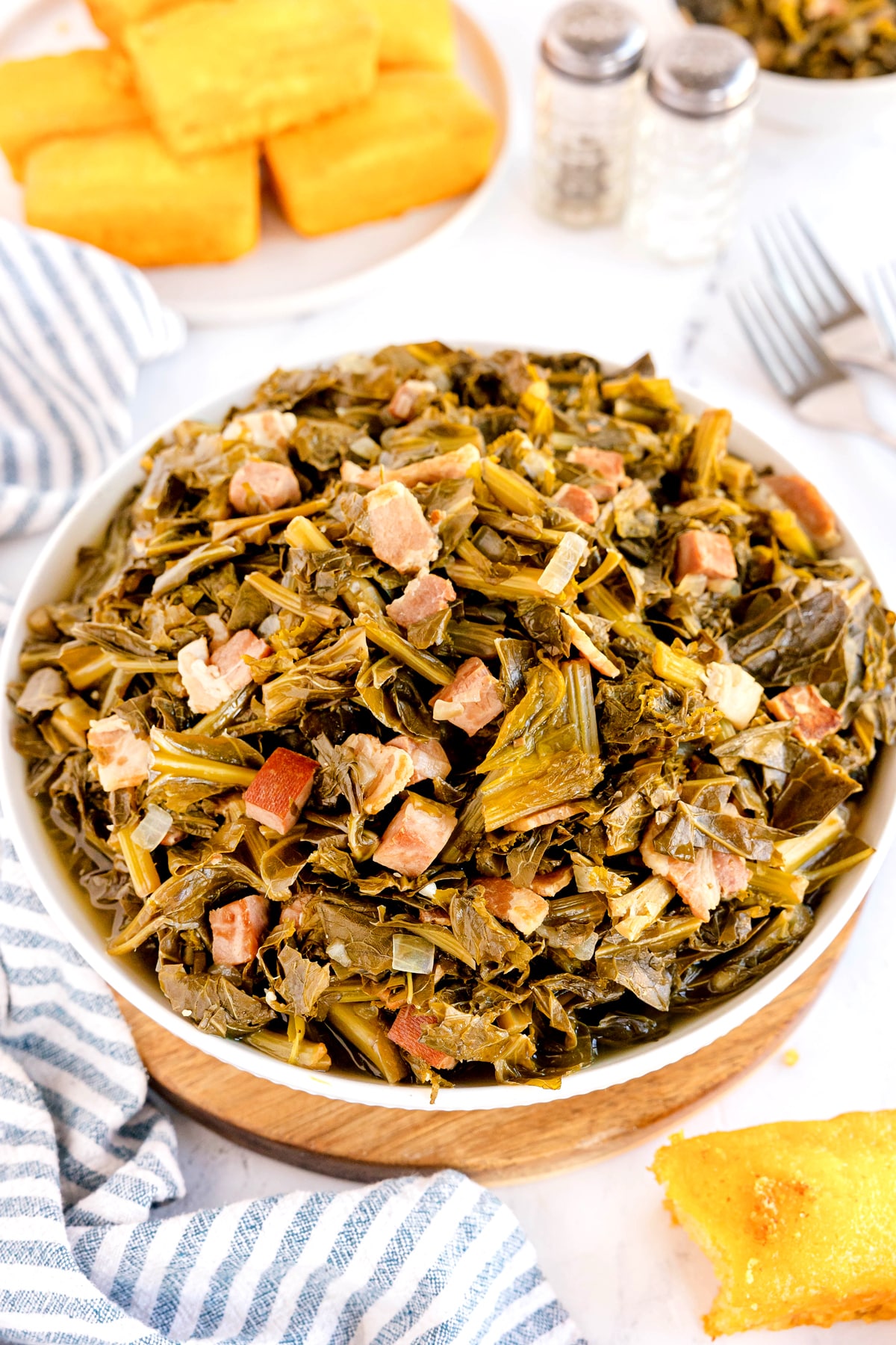 A bowl of cooked Southern Collard Greens mixed with chopped pieces of bacon, served on a table with cornbread, salt and pepper shakers, and a striped napkin nearby.