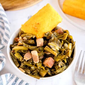 A bowl of cooked Southern Collard Greens with diced pork, topped with a piece of cornbread. A fork and a striped napkin are nearby.