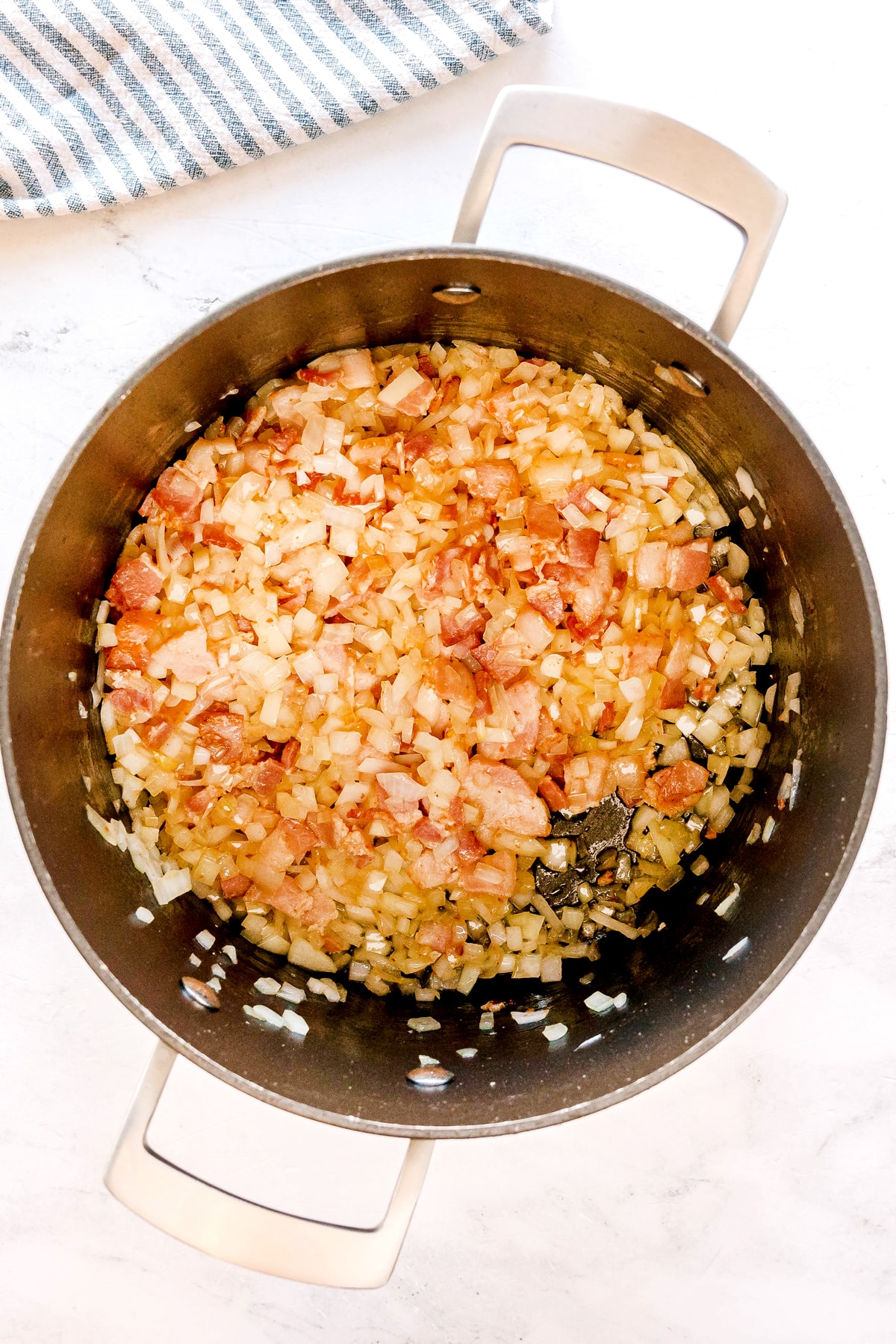 Next step in preparing Southern Collard Greens is to sauté chopped onions and bacon in a large pot on a light-colored countertop, with a striped towel partially visible in the corner.