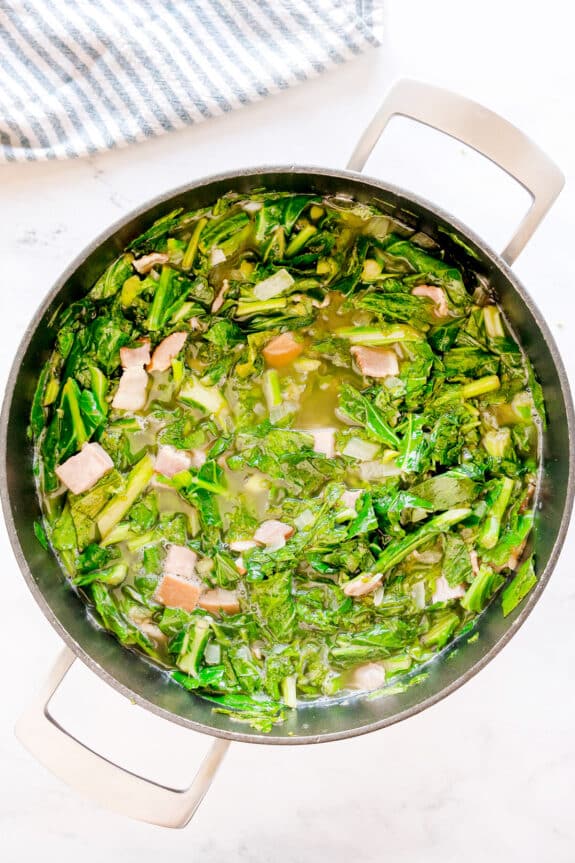A pot of Southern Collard Greens consisting of simmering greens with pieces of ham in collard greens, apple cider vinegar, sugar, salt, and black pepper and broth, viewed from above, on a white surface with a striped cloth nearby.