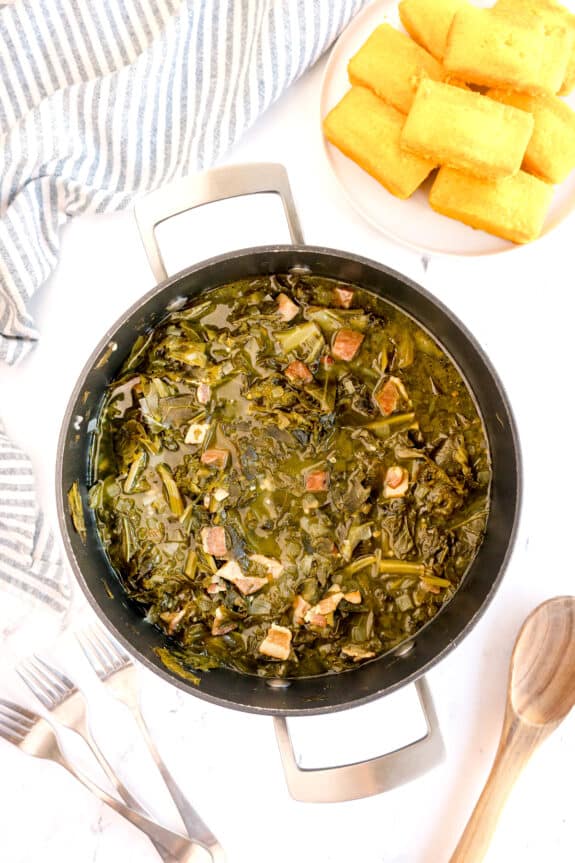A pot of cooked Southern Collard Greens with ham pieces, next to a plate of cornbread, striped towel, wooden spoon, and forks on a white surface.