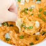 A hand scoops a chip into creamy, high protein buffalo chicken dip topped with chopped green onions and blue cheese crumbles.