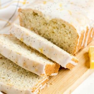 A loaf of Lemon Poppy Seed Bread with white icing is sliced on a wooden cutting board, highlighting its moist texture and zesty flavor.
