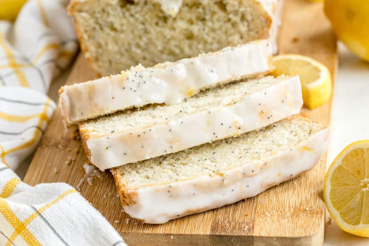 Sliced Lemon Poppy Seed Bread with white icing sits on a wooden board, surrounded by fresh lemon slices and a striped towel.