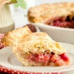 A slice of fruit pie with a golden, flaky crust and vibrant red filling sits on a white plate—the perfect showcase for any classic rhubarb pie recipe—with the rest of the pie visible in the background.