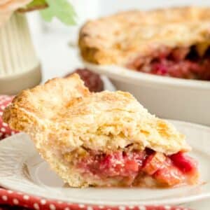 A slice of fruit pie with a golden, flaky crust and vibrant red filling sits on a white plate—the perfect showcase for any classic rhubarb pie recipe—with the rest of the pie visible in the background.