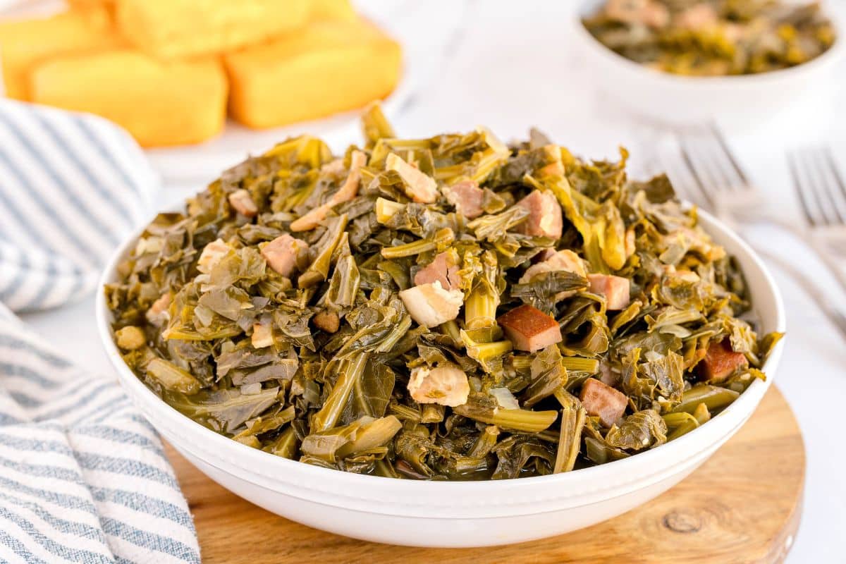 A bowl of cooked collard greens with pieces of meat, set on a wooden board with cornbread and utensils in the background, highlights a classic Southern Collard Greens Recipe.