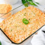 A glass baking dish filled with a cheesy Jalapeno Corn Dip, topped with two sliced jalapeños, sits on a white surface with shredded cheese and peppers nearby.