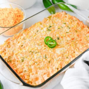A glass baking dish filled with a cheesy Jalapeno Corn Dip, topped with two sliced jalapeños, sits on a white surface with shredded cheese and peppers nearby.