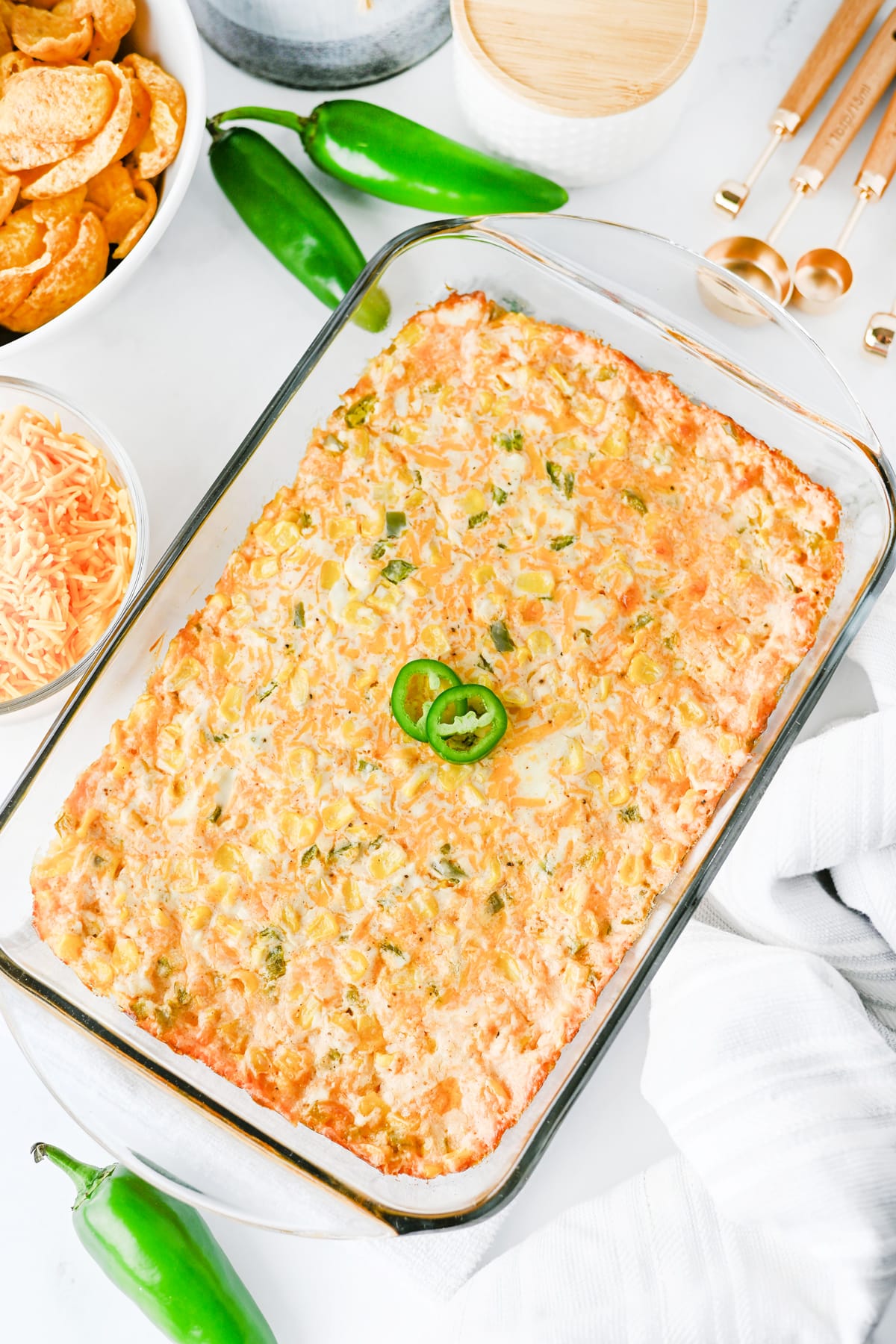 A glass baking dish filled with cheesy Jalapeno Corn Dip. Surrounding the dish are corn chips, shredded cheese, jalapeños, and measuring spoons.