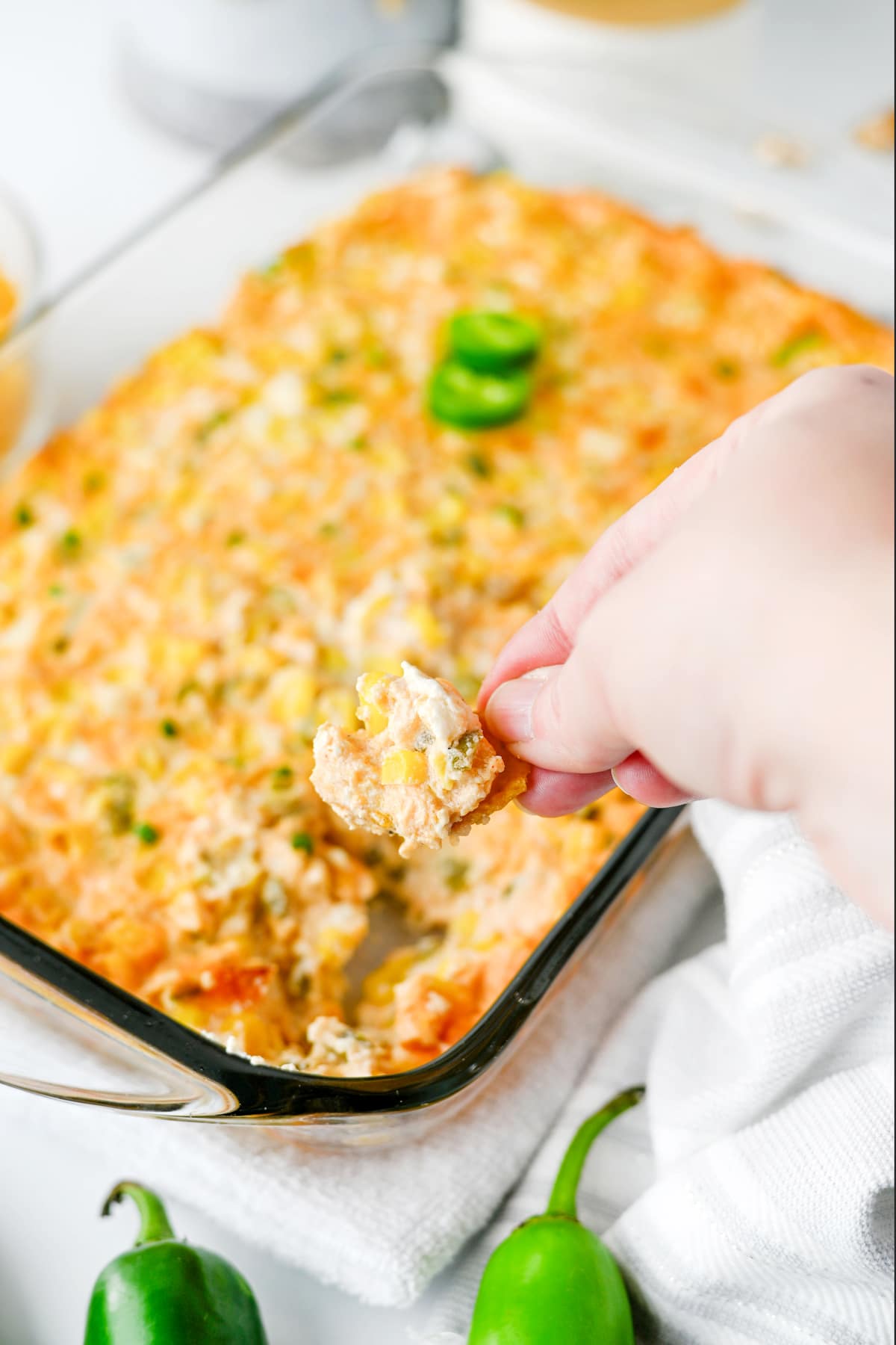 A hand holds a chip with Jalapeno Corn Dip above a glass baking dish containing the dip. Two green jalapeños and a white towel are nearby.