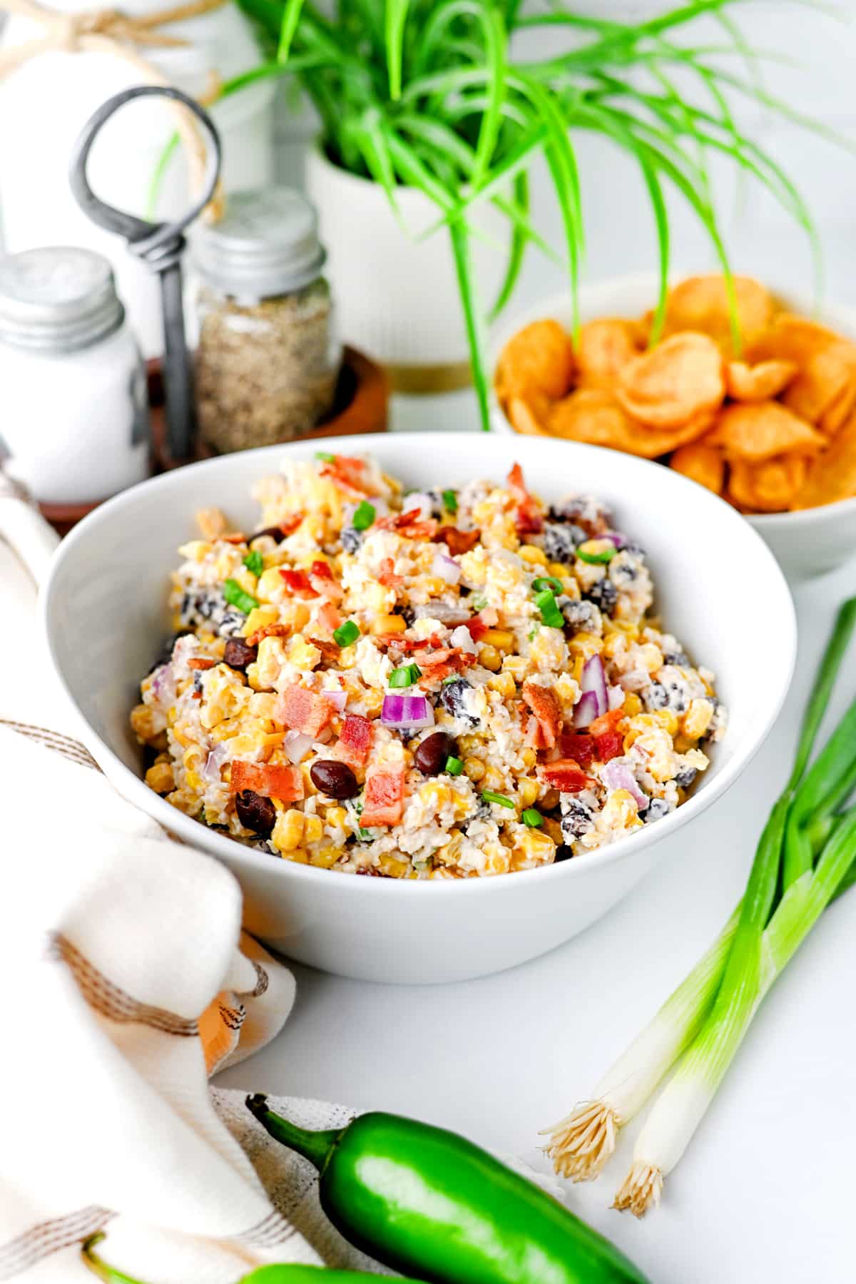 A white bowl of Cowboy Corn Dip with bacon, black beans, and red onions sits on a table near green onions, jalapeño, pork rinds, salt and pepper shakers, and a potted plant.