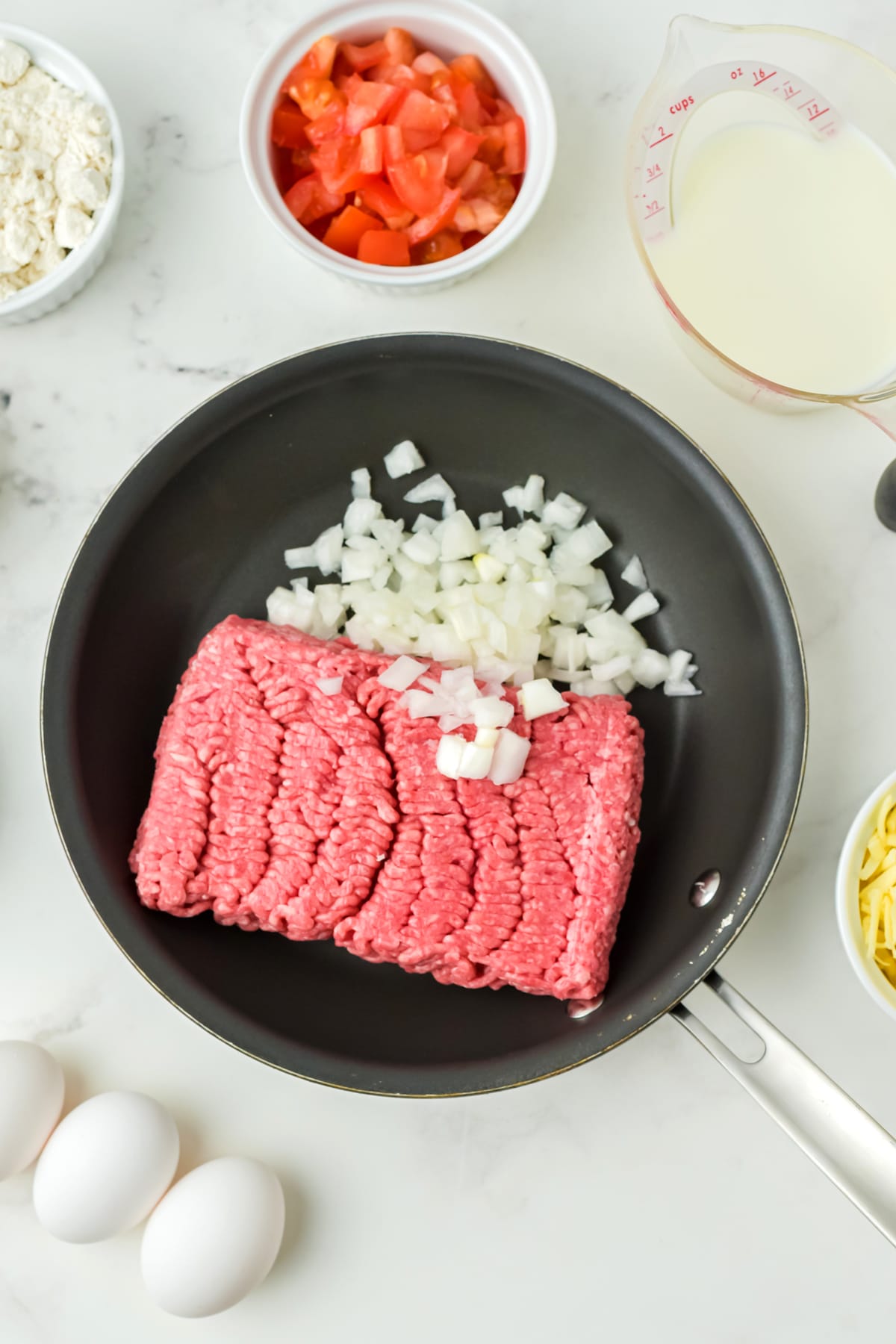 A frying pan with raw ground beef and chopped onions for Taco Recipe, surrounded by bowls of flour, diced tomatoes, shredded cheese, eggs, and a measuring cup of milk.