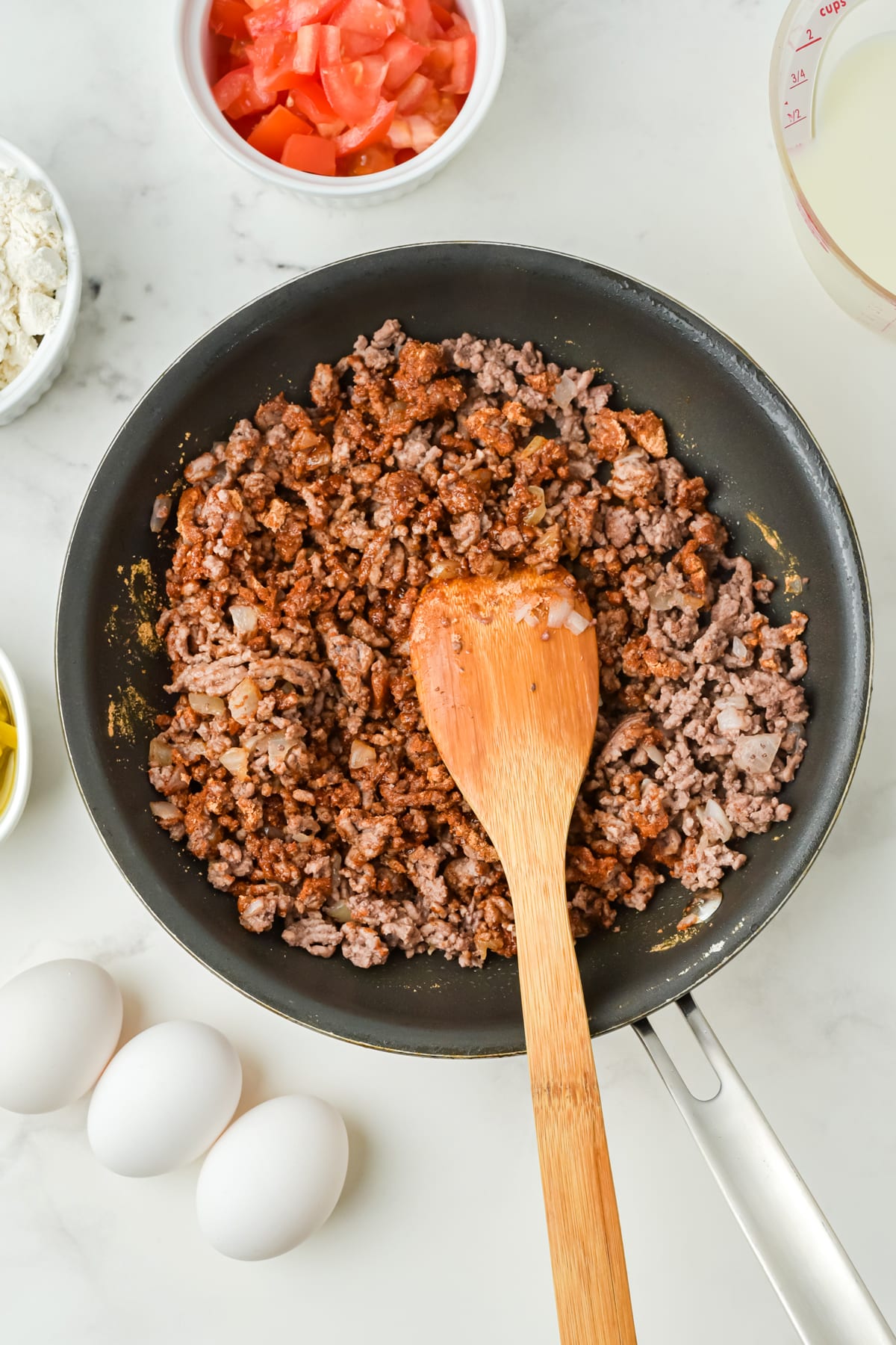 Ground beef for Taco Pie is browned in a black skillet with a wooden spoon, surrounded by eggs, diced tomatoes, added with taco seasoning, flour, and a measuring cup of liquid on a white countertop.