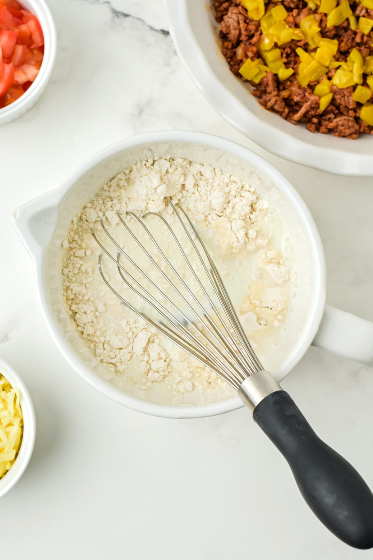 A mixing bowl with Bisquick and liquid being whisked, surrounded by bowls of shredded cheese, diced tomatoes, and cooked ground meat with chopped peppers. This is one of the steps in preparing Taco Pie Recipe
