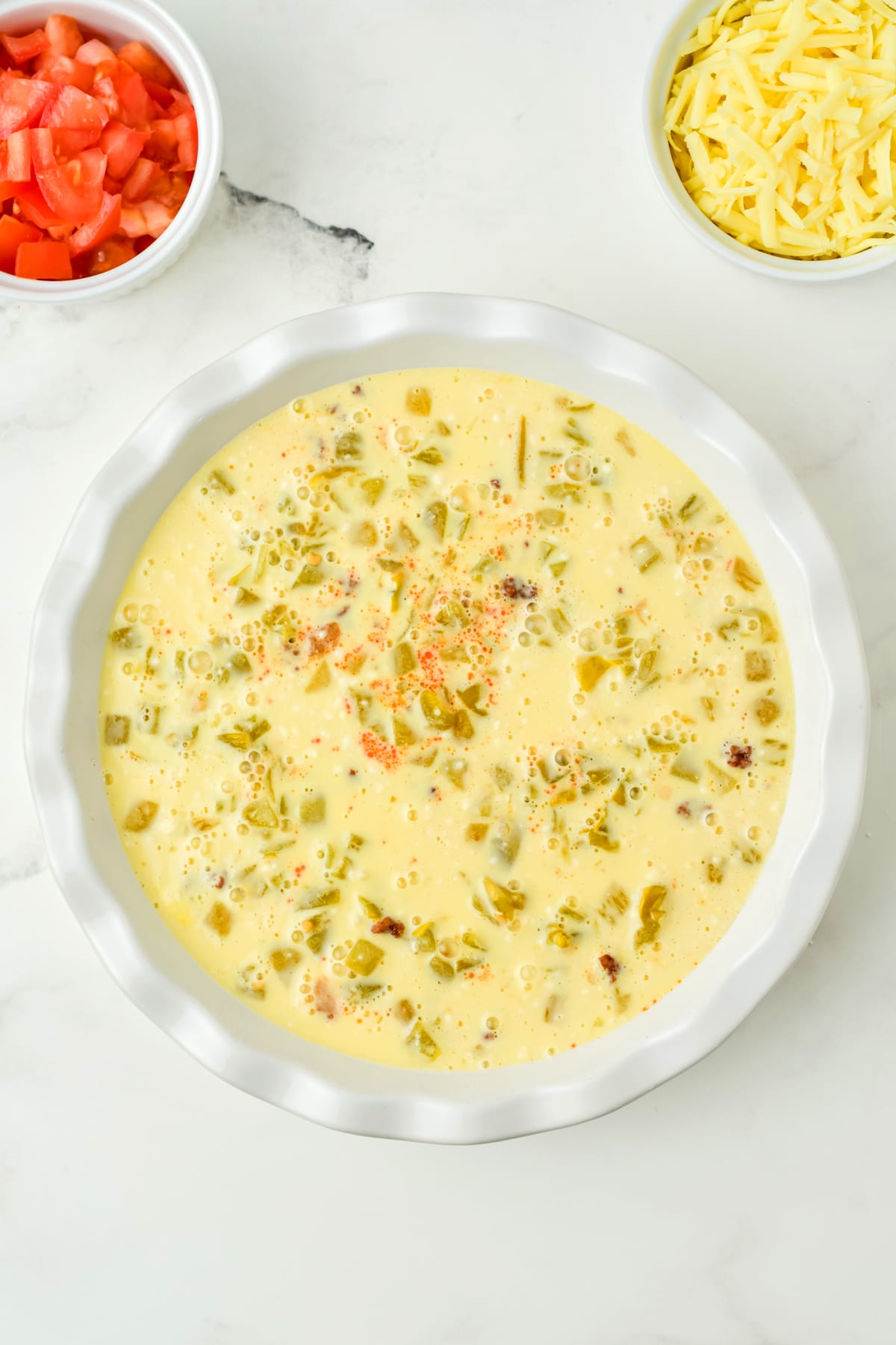 A white pie dish filled with an Bisquick, eggs, and milk mixture topped with chopped green chilies, next to small bowls of diced tomatoes and shredded cheese on a white surface. This is for Taco Pie recipe.