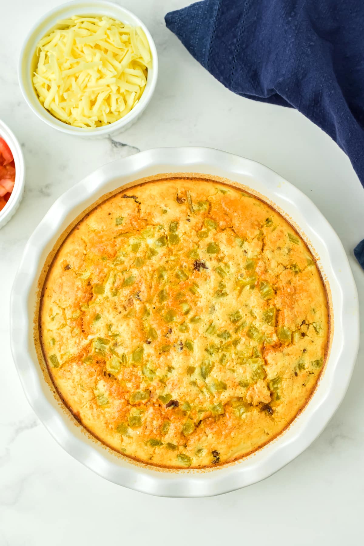 A baked casserole Taco Pie with a golden top, surrounded by small bowls of shredded cheese and diced tomatoes on a white surface.