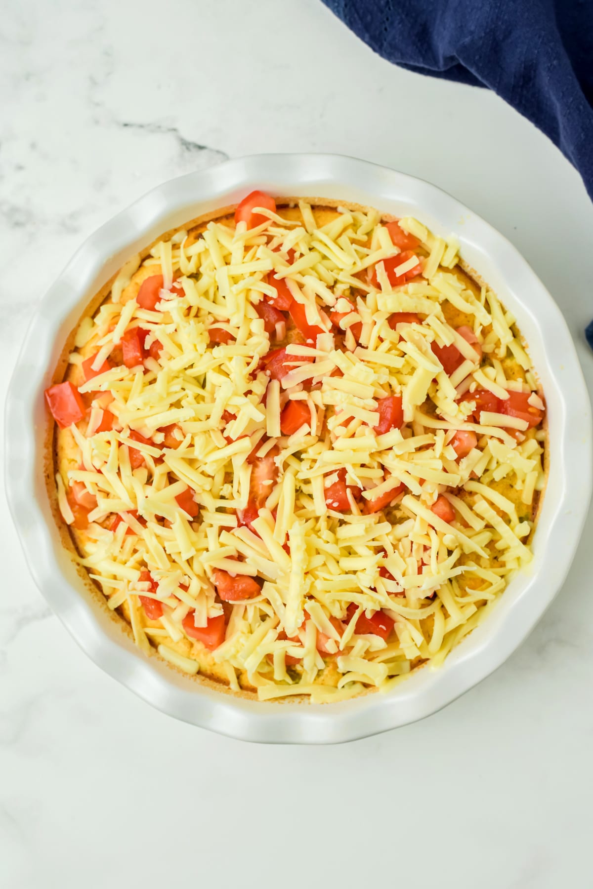 A round baking dish of Taco Pie, filled with an unbaked casserole topped with shredded cheese and chopped tomatoes, on a marble countertop.