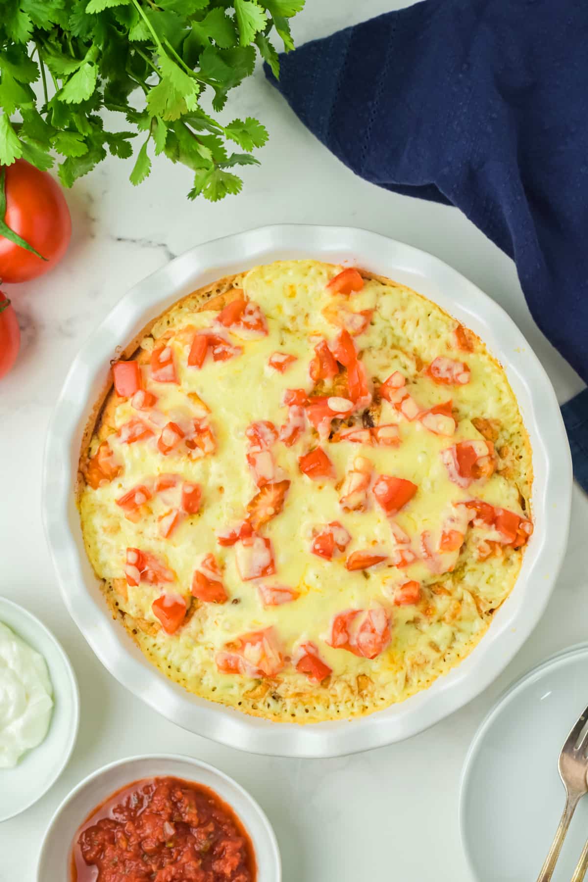 Freshly-baked Taco Pie recipe topped with melted cheese and diced tomatoes, surrounded by bowls of salsa, sour cream, fresh cilantro, a tomato, and a blue napkin on a white surface.