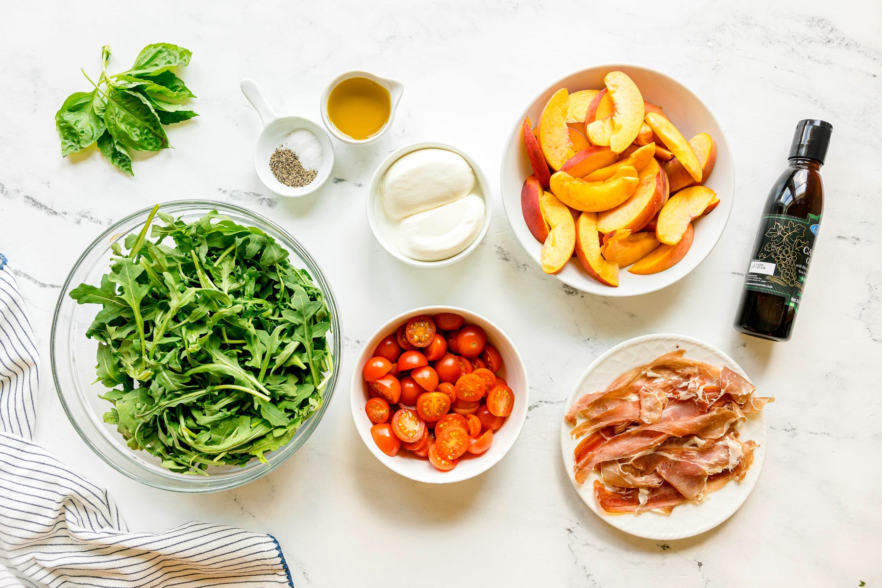 Bowls of arugula, cherry tomatoes, peach slices, prosciutto, and jars of olive oil, burrata cheese, fresh basil leaves, balsamic glaze, kosher salt, and ground black pepper are arranged on a white countertop—perfect ingredients for assembling a refreshing Burrata Peach Salad.