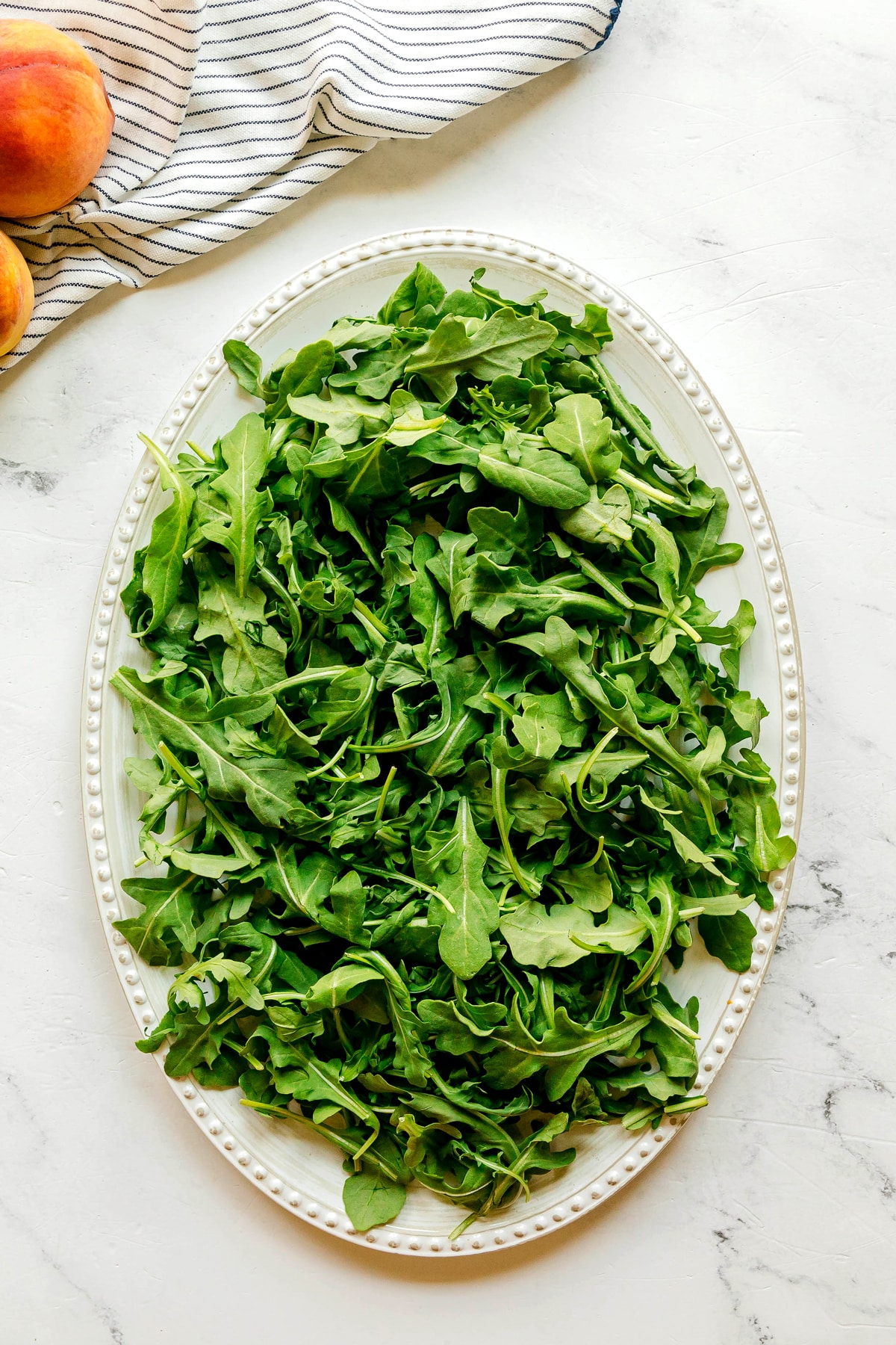 A white oval plate filled with fresh arugula leaves sits on a marble surface, hinting at a delicious Burrata Peach Salad, with a striped cloth and ripe peaches partially visible in the background.