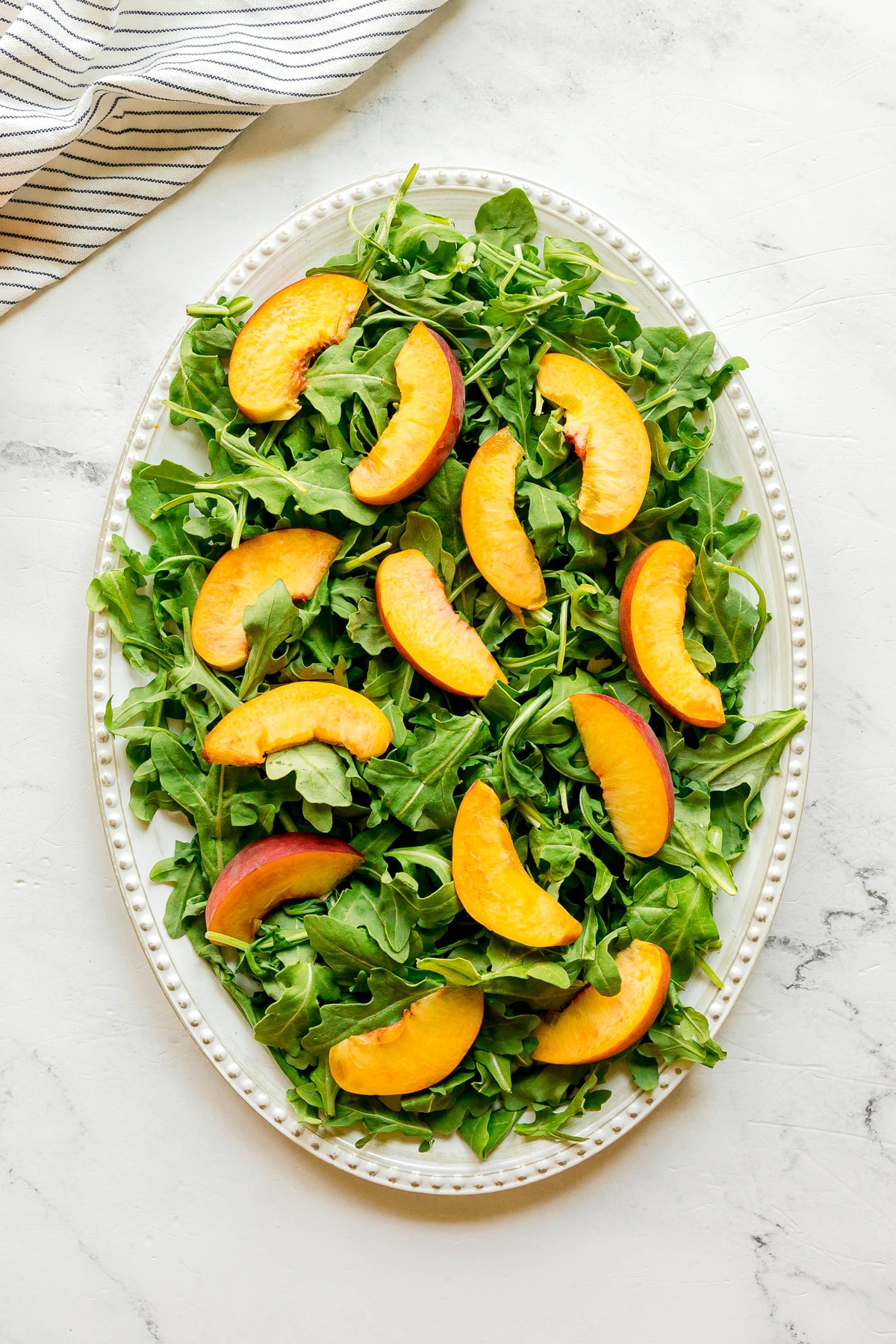 Oval platter with arugula leaves topped with peach slices, creating a vibrant Burrata Peach Salad, placed on a white marble surface next to a striped cloth.