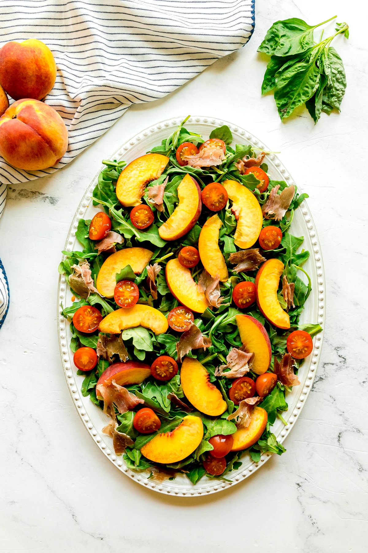Oval platter with arugula, sliced peaches, cherry tomatoes, and prosciutto on a marble surface; striped cloth, basil, whole peaches, and creamy burrata evoke the freshness of a Burrata Peach Salad.