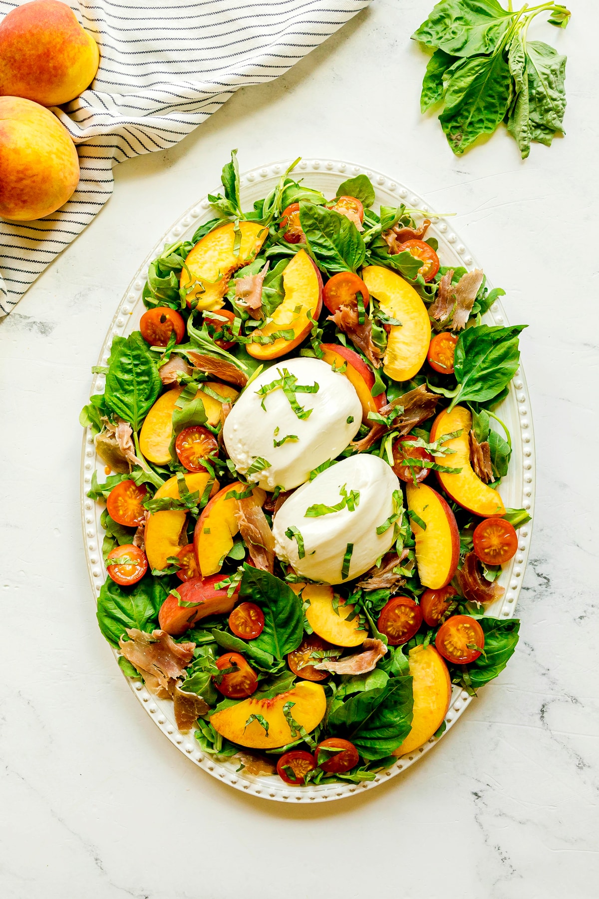 An oval platter with burrata cheese, peach slices, cherry tomatoes, prosciutto, and fresh basil on a bed of greens sits on a white surface next to a striped napkin, peaches, and basil leaves. This is a perfect plate of Burrata Peach Salad